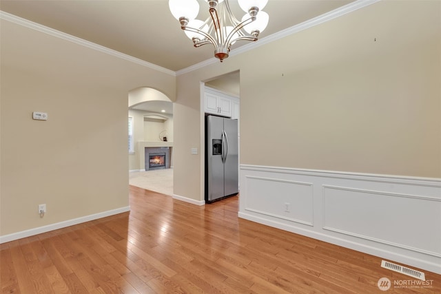 spare room with visible vents, a glass covered fireplace, a wainscoted wall, light wood-style flooring, and crown molding