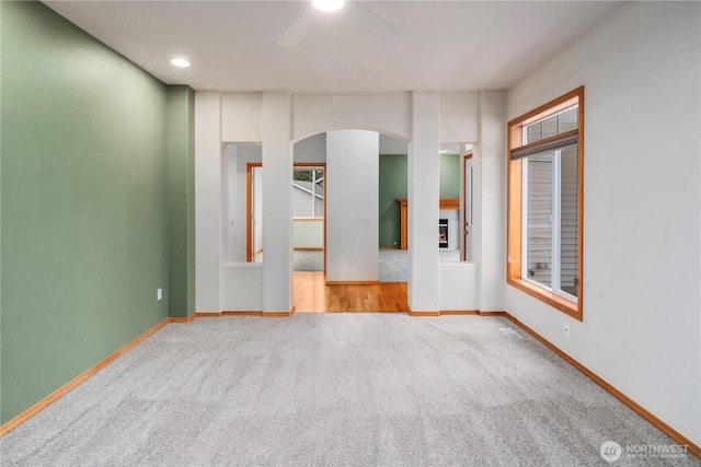 carpeted empty room with arched walkways, a warm lit fireplace, ceiling fan, and baseboards