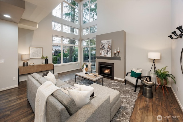 living area featuring a towering ceiling, baseboards, dark wood-type flooring, and a glass covered fireplace