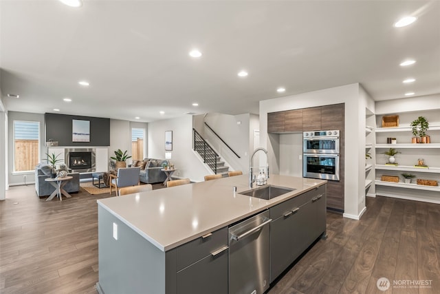 kitchen with a sink, appliances with stainless steel finishes, dark wood-style floors, a glass covered fireplace, and a kitchen island with sink