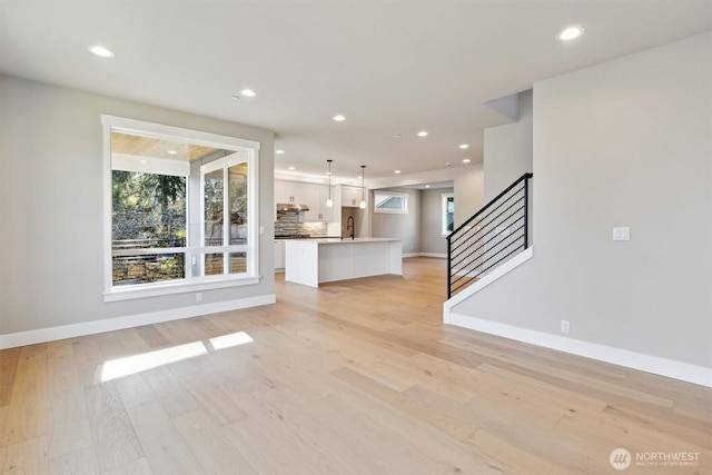 unfurnished living room featuring baseboards, light wood-style flooring, stairs, a sink, and recessed lighting