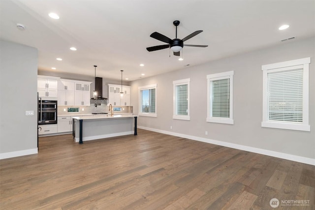 kitchen with light countertops, open floor plan, white cabinets, an island with sink, and wall chimney exhaust hood