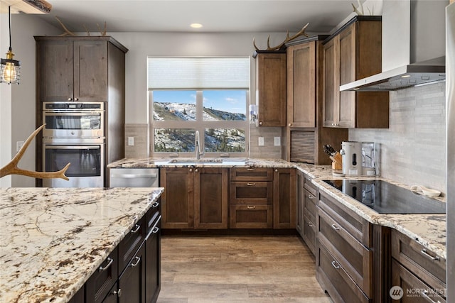 kitchen with tasteful backsplash, wall chimney range hood, light wood-type flooring, stainless steel appliances, and a sink