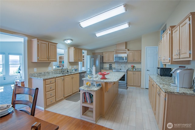 kitchen with a center island with sink, stainless steel appliances, light brown cabinetry, vaulted ceiling, and a sink