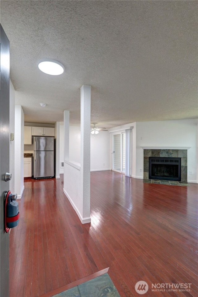 unfurnished living room with ceiling fan, a textured ceiling, a tile fireplace, dark wood-type flooring, and baseboards