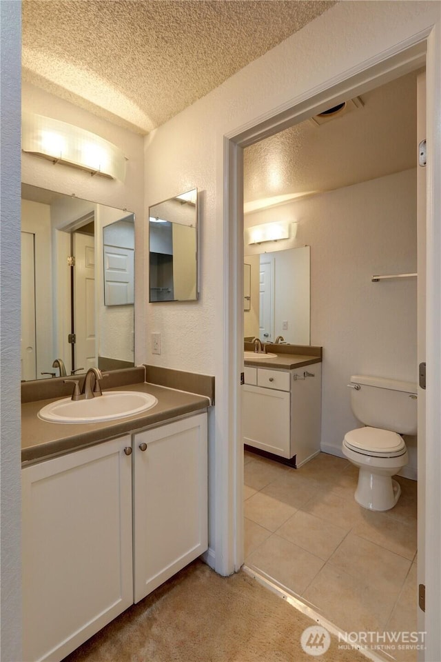 full bath featuring toilet, tile patterned floors, a sink, a textured ceiling, and two vanities