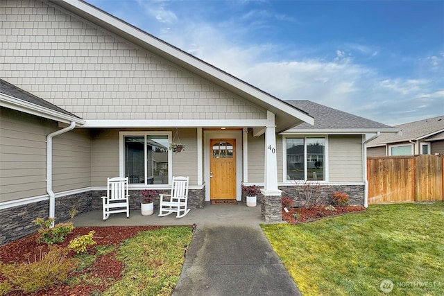 view of front of house featuring roof with shingles, covered porch, fence, stone siding, and a front lawn