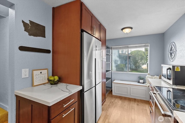 kitchen featuring stainless steel appliances, a textured wall, brown cabinetry, light stone countertops, and light wood-type flooring