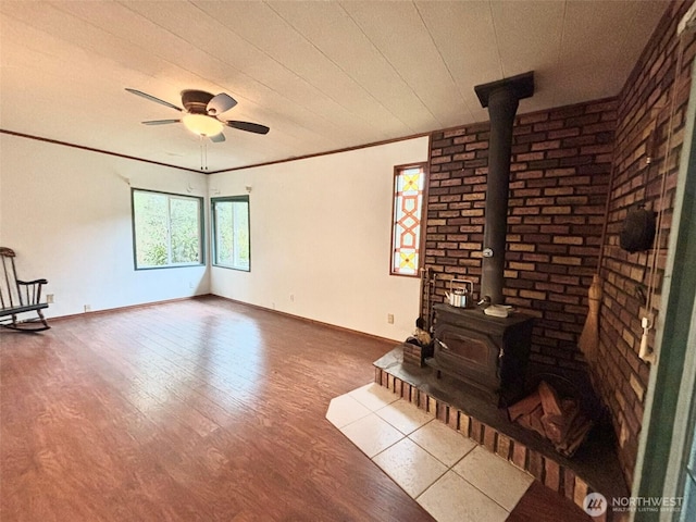 unfurnished living room featuring crown molding, a wood stove, ceiling fan, wood finished floors, and baseboards