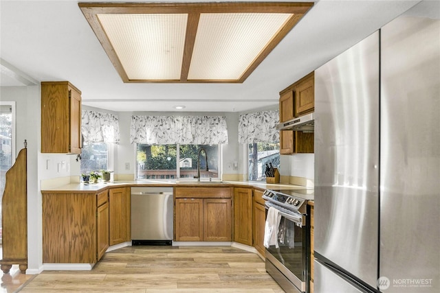 kitchen with under cabinet range hood, stainless steel appliances, a sink, light countertops, and brown cabinets