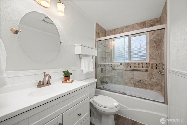 full bathroom featuring a wainscoted wall, bath / shower combo with glass door, vanity, and toilet