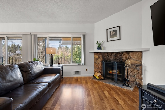 living room featuring baseboards, visible vents, wood finished floors, a textured ceiling, and a stone fireplace