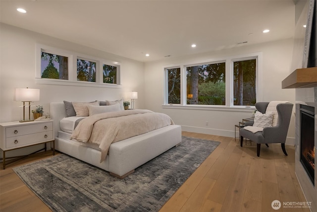 bedroom with baseboards, light wood-type flooring, a glass covered fireplace, and recessed lighting