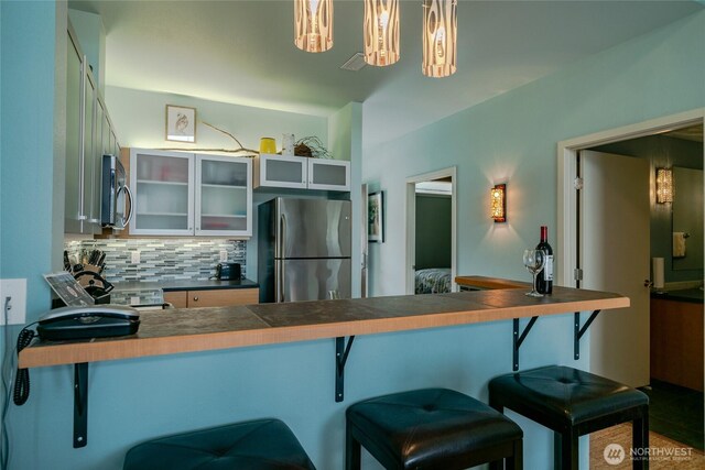 kitchen with decorative backsplash, appliances with stainless steel finishes, glass insert cabinets, a peninsula, and a kitchen bar