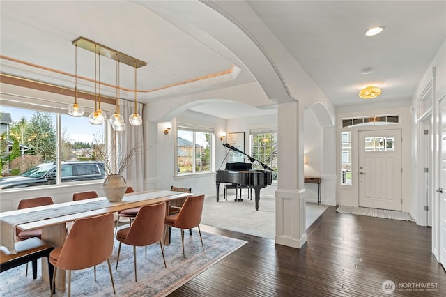 dining space with dark wood-style floors, decorative columns, arched walkways, ornamental molding, and a raised ceiling