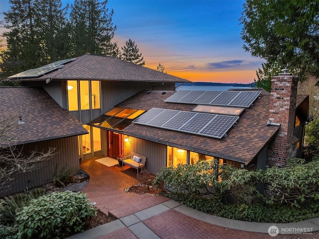 back of house at dusk with solar panels, roof with shingles, and a chimney