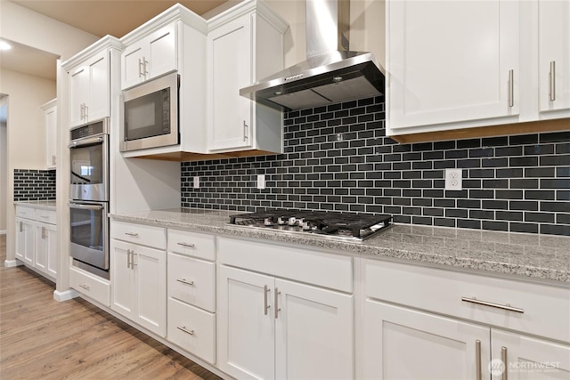 kitchen featuring light wood-type flooring, appliances with stainless steel finishes, white cabinets, wall chimney range hood, and decorative backsplash