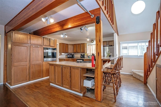 kitchen with fridge, brown cabinetry, beam ceiling, light countertops, and stainless steel oven