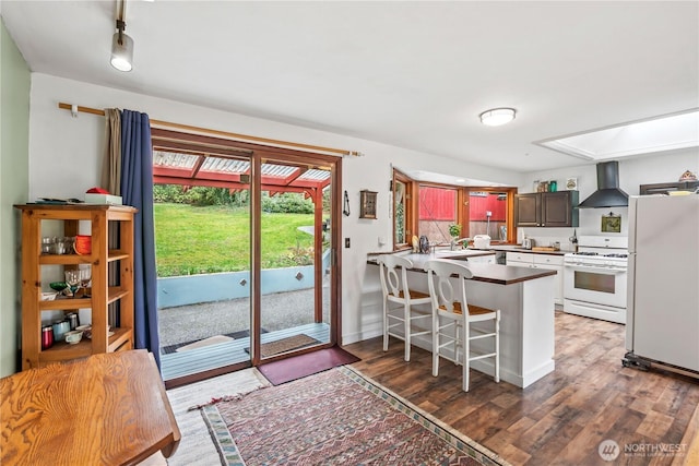 kitchen with a sink, wood finished floors, white appliances, a peninsula, and wall chimney exhaust hood