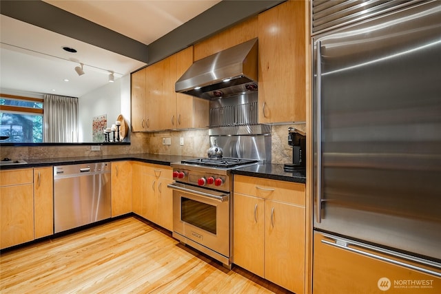 kitchen with tasteful backsplash, wall chimney range hood, dark stone counters, light wood-type flooring, and high end appliances