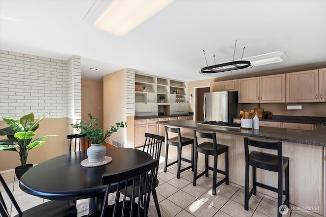 kitchen featuring light tile patterned floors, dark countertops, light brown cabinetry, appliances with stainless steel finishes, and a kitchen bar