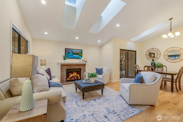 living area featuring light wood-type flooring, vaulted ceiling with skylight, a notable chandelier, and a brick fireplace