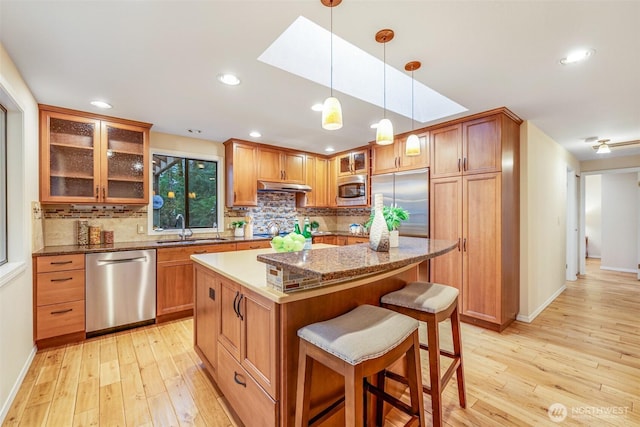 kitchen with light wood finished floors, a kitchen island, under cabinet range hood, appliances with stainless steel finishes, and a sink