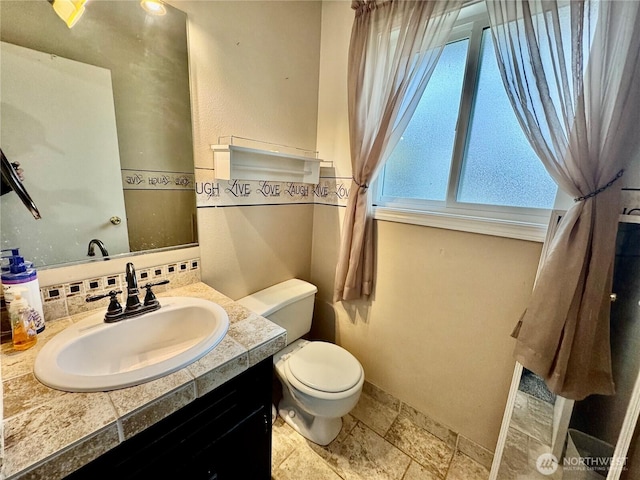 half bathroom featuring vanity, stone tile floors, baseboards, decorative backsplash, and toilet