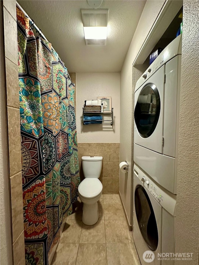 laundry area with a wainscoted wall, stacked washer / drying machine, tile patterned floors, a textured ceiling, and tile walls