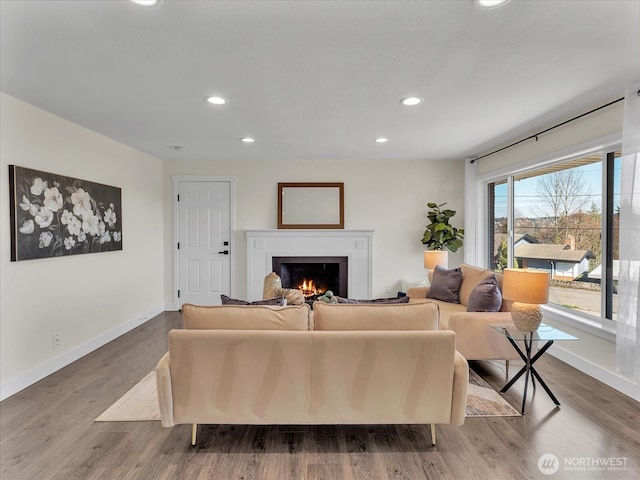 living room featuring recessed lighting, a warm lit fireplace, baseboards, and wood finished floors