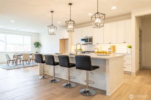 kitchen with light wood finished floors, stainless steel microwave, a center island with sink, and backsplash