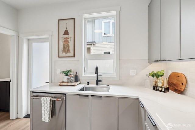 kitchen featuring gray cabinetry, a sink, light wood finished floors, decorative backsplash, and dishwasher