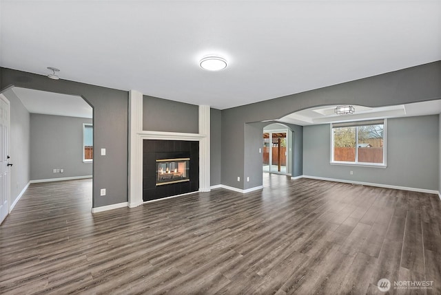 unfurnished living room with arched walkways, dark wood-type flooring, a tile fireplace, and baseboards