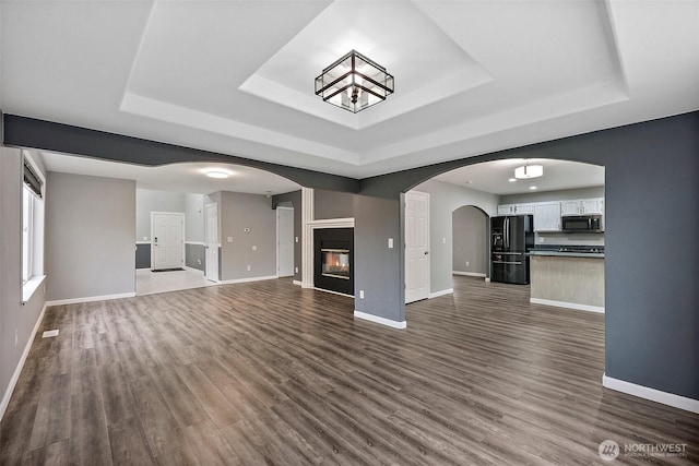 unfurnished living room featuring arched walkways, dark wood-type flooring, a raised ceiling, and a glass covered fireplace