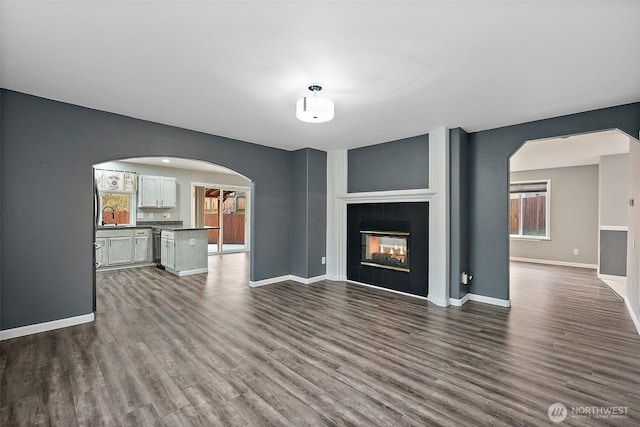 unfurnished living room with arched walkways, a fireplace, baseboards, a wealth of natural light, and dark wood finished floors