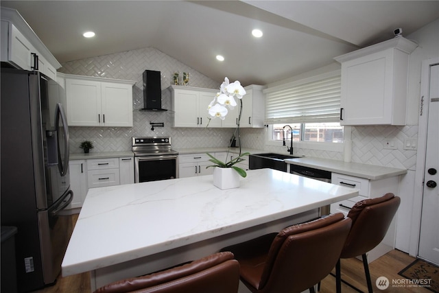 kitchen with a breakfast bar area, a sink, vaulted ceiling, appliances with stainless steel finishes, and wall chimney exhaust hood