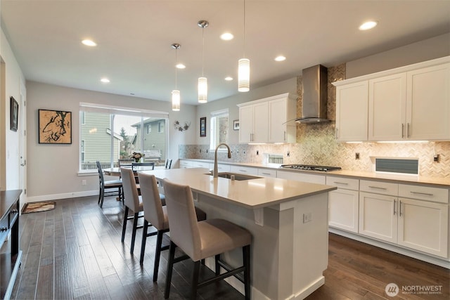 kitchen featuring a center island with sink, a sink, wall chimney range hood, tasteful backsplash, and light countertops