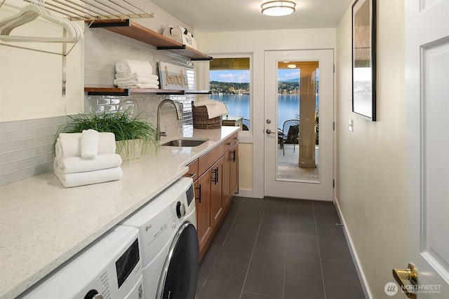 clothes washing area featuring a water view, dark tile patterned flooring, a sink, baseboards, and washing machine and clothes dryer