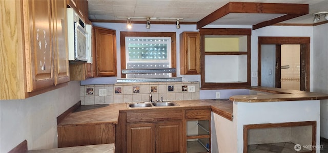 kitchen featuring tile countertops, backsplash, white microwave, brown cabinetry, and a sink