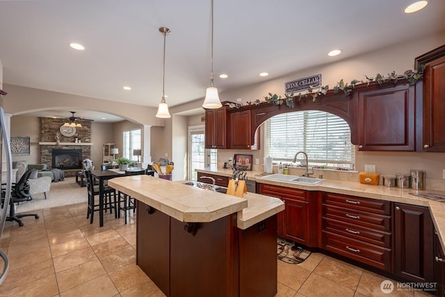 kitchen with a sink, light countertops, a stone fireplace, a ceiling fan, and reddish brown cabinets