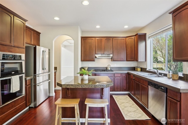 kitchen with appliances with stainless steel finishes, a sink, a kitchen island, a breakfast bar, and dark wood-type flooring