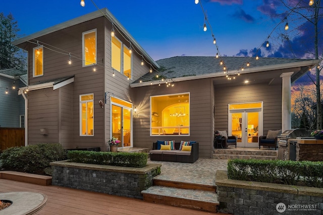 back of property at dusk featuring a deck, french doors, roof with shingles, exterior kitchen, and outdoor lounge area