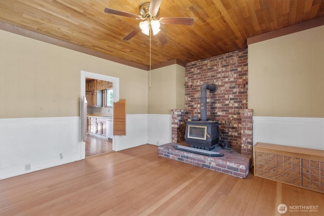 unfurnished living room featuring a wainscoted wall, a wood stove, ceiling fan, wood finished floors, and wooden ceiling