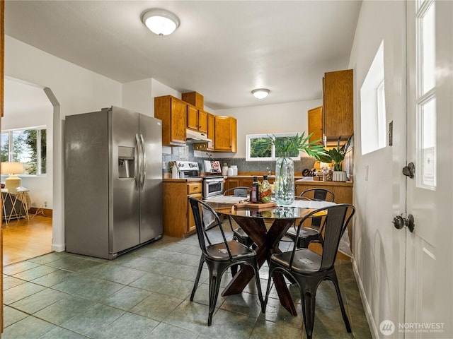 kitchen featuring under cabinet range hood, stainless steel appliances, arched walkways, brown cabinetry, and decorative backsplash