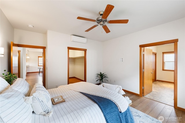 bedroom with a spacious closet, baseboards, an AC wall unit, wood finished floors, and a ceiling fan