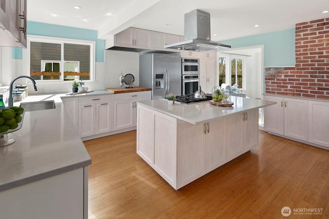 kitchen featuring island exhaust hood, a sink, appliances with stainless steel finishes, light wood finished floors, and light countertops