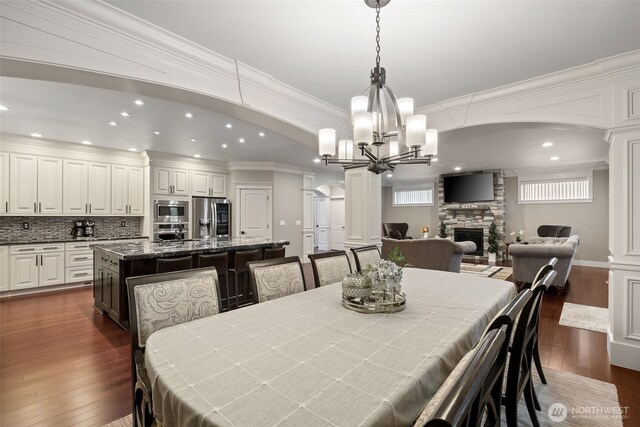 dining room with arched walkways, a stone fireplace, dark wood finished floors, and ornamental molding