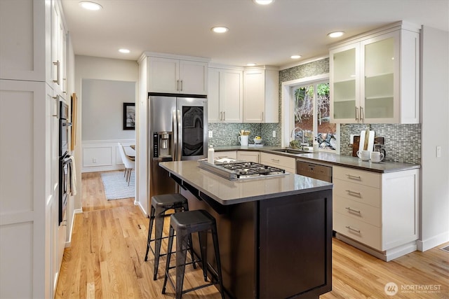 kitchen with a sink, glass insert cabinets, light wood-style floors, appliances with stainless steel finishes, and a kitchen breakfast bar