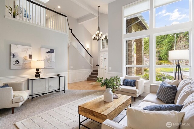 carpeted living area featuring a wainscoted wall, beam ceiling, a high ceiling, an inviting chandelier, and stairs