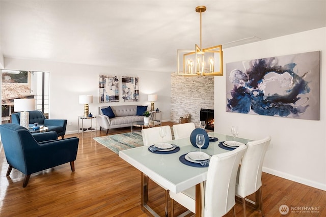 dining space featuring baseboards, a notable chandelier, a stone fireplace, and wood finished floors